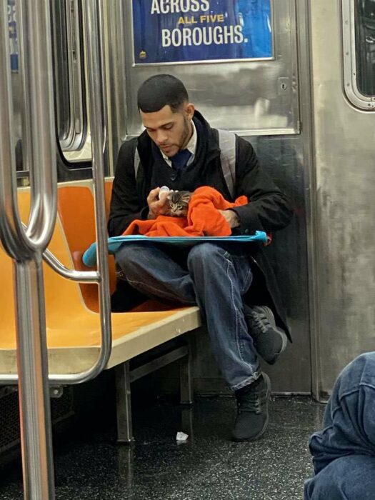Hombre sentado en el metro con saco negro, camisa blanca, corbata negra y jeans, alimenta a un gatito café con rayas negras mientras lo envuelve con una manta naranja