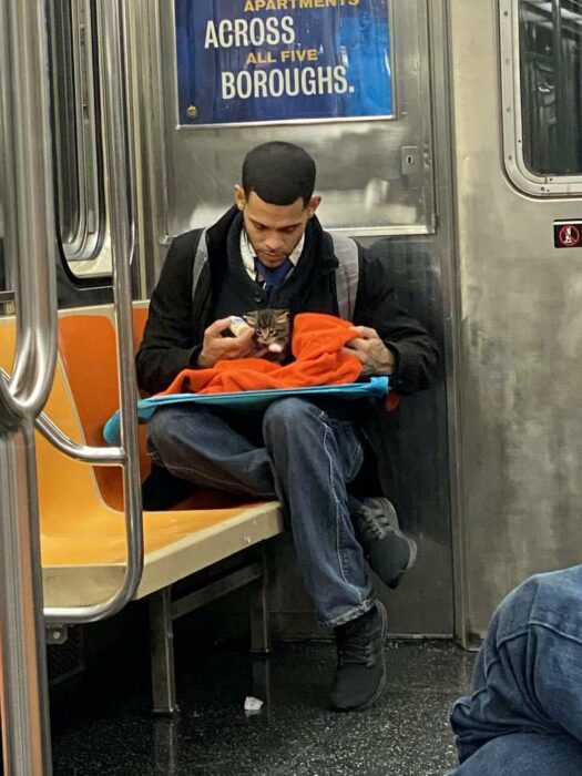 Hombre sentado en el metro con saco negro, camisa blanca, corbata negra y jeans, alimenta a un gatito café con rayas negras mientras lo envuelve con una manta naranja