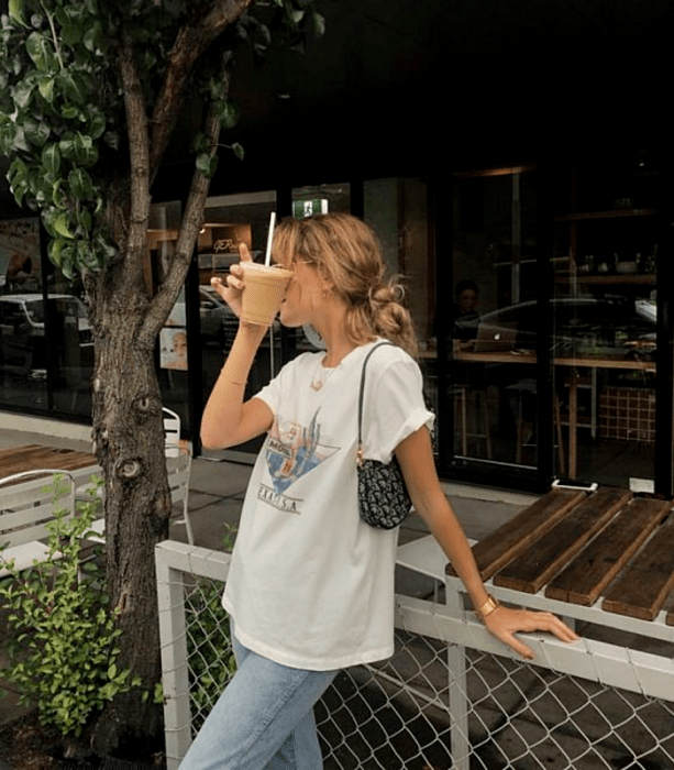 chica rubia usando lentes, camiseta blanca estampada, jeans holgados y bolso de mano negro con blanco