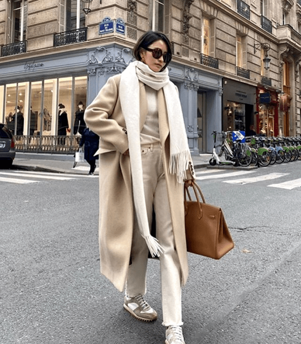 chica de cabello castaño usando lentes de sol, bufanda blanca, top blanco, abrigo largo beige, pantalones blancos, tenis deportivos beige, bolso café claro de piel