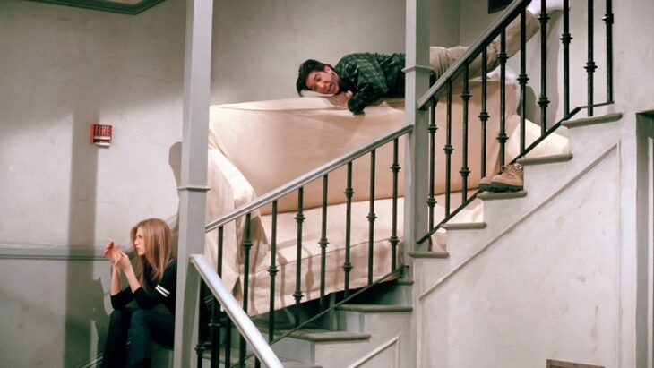 Rachel y Ross de Friends en las escaleras de un edificio tratando de bajar un sillón beige