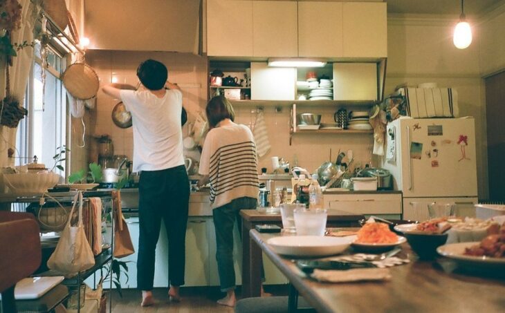 Hombre de camiseta blanca y pantalón negro, y mujer bajita de cabello corto con pantalones de mezclilla y suéter blanco con rayas negras cocinando