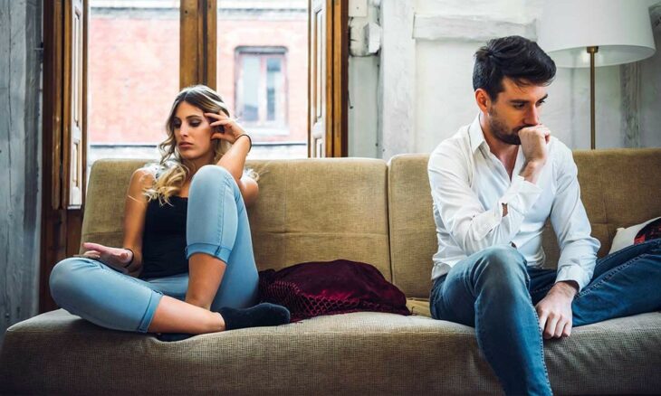 Mujer rubia con cabello ondulado, blusa de tirantes negra y jeans claros, haciendo gesto de disgusto; junto a un hombre con barba, camisa blanca y jeans oscuros, pensativo en un sillón beige