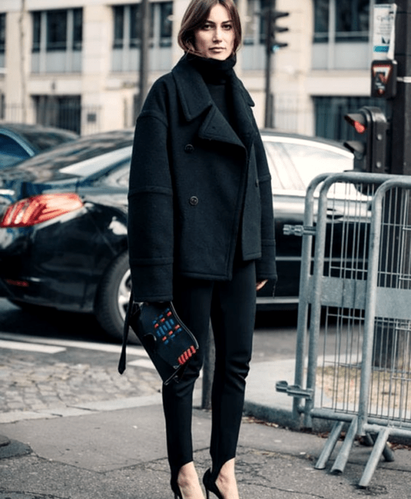 Chica de cabello castaño usando un suéter de cuello alto negro, abrigo grande negro, leggings con estribos negros, zapatos de tacón y bolso de mano negro con vistas rojas y azules