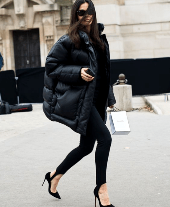 Chica de cabello castaño usando lentes de sol, abrigo puff jacket oversized, leggings negros con estribos, zapatos de tacón y suéter negro largo