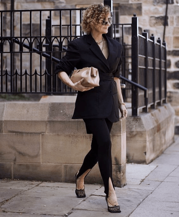 Chica de cabello chino rubio usando lentes de sol, blazer oversized negro, leggings negros con estribos y zapatos de tacón semitransparentes, bolso de mano beige