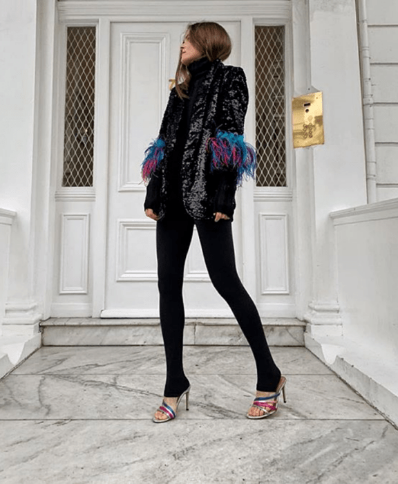 Chica de cabello claro usando un top negro de cuello alto, blazer negro de brillantina con plumas azules y rosas en las mangas, leggings negros con estribos y sandalias de tacón de colores