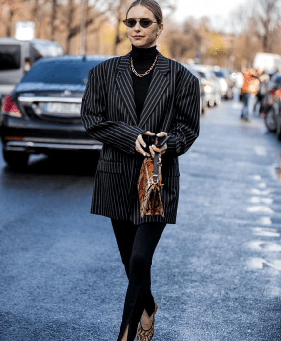 Chica rubia con lentes de sol, top negro de cuello alto, blazer oversized negro de líneas negras, leggings negros con estribos, sandalias de tacón negras, bolso de mano café con animal print