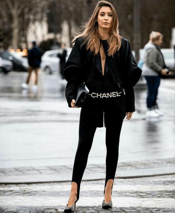 Chica de cabello claro usando una blusa negra de mangas abombadas largas, leggings negros con estribos, cinturón de chanel, bolso negro de mano y zapatos negros de tacón con aplicaciones de brillantes en la punta