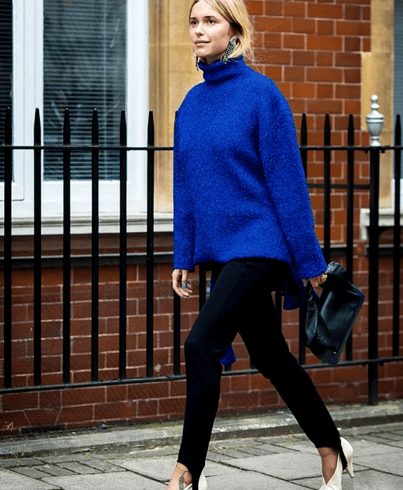 Chica rubia usando un suéter azul de cuello alto, leggings negros con estribos, zapatos blancos de tacón y bolso negro de mano