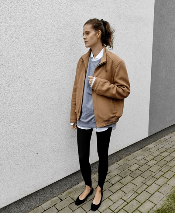 Chica de cabello castaño usando una camisa de vestir blanca, sudadera gris, chamarra café claro, leggings negros con estribos, flats negros