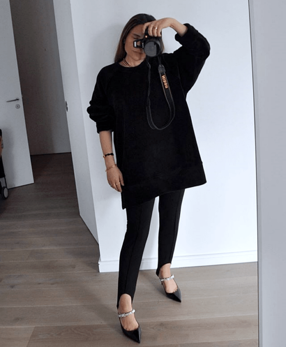 Chica de cabello castaño largo usando un suéter negro oversized, leggings negros de estribos, zapatos negros de tacón con correas con brillantes