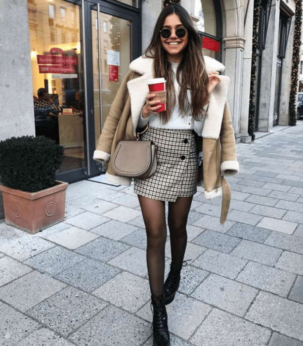 chica de cabello largo usando gafas de sol, camiseta blanca, chaqueta de borreguito, medias oscuras, falda de cuadros blanca y negra, y botines