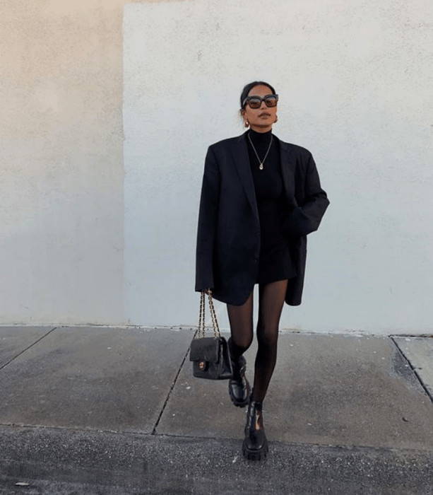 chica con cabello oscuro, gafas de sol, camiseta de manga larga, blazer oversize, medias negras, minifalda negra y botines negros