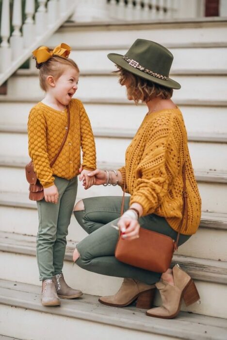 Madre e hija sonriendo en escalones blancos ambas con atuendo iguales de pantalón verde oliva, botines beige, suéter tejido amarillo mostaza, bolso camel mientras que la niña usa un moño grande amarillo y la madre un sombrero verde olivo