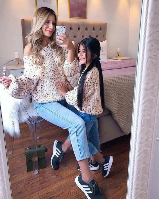 Madre de cabello rubio e hija pequeña de cabello largo y lacio se toman selfie frente al espejo recargadas en la cama mientras usan jeans claros, tenis negros con rayas blancas y un suéter claro con bolitas negras