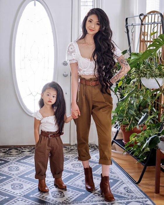 Madre e hija posando en la entrada de su casa con paredes blancas y plantas verdes de decoración a un costado, ambas vistiendo pantalón café, botines cafés, blusa blanca de encaje y cabello ondulado largo peinado hacia un costado