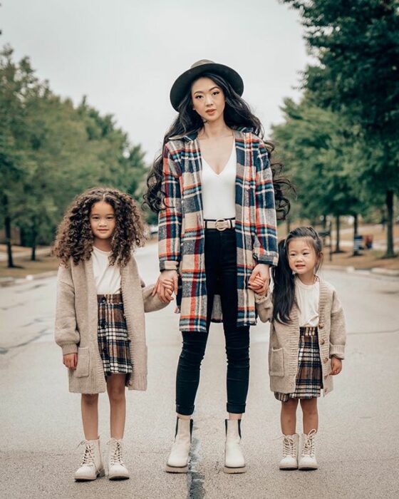 Madre asiática y delgada de cabello largo ondulado con sombrero gris vistiendo blusa blanca, pantalón negro, botines blancos y saco de cuadros en color beig, rojo y azul, sostiene la manos de sus dos hijas que viste botines beige, falda de cuadros café con blanco, blusa blanca y cardigan largo en tonos beige
