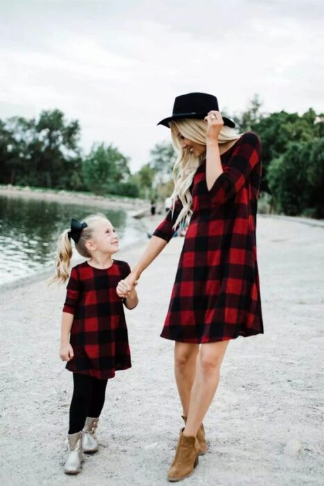 Madre e hija tomadas de la mano cerca de un lago las dos con vestido rojo con cuadros negros, la madre usa botines cafés y sombrero negro con el cabello largo, rubio y ondulado y la niña usa mallones negros con botas plateadas con coleta alta de lado y un moño negro grande
