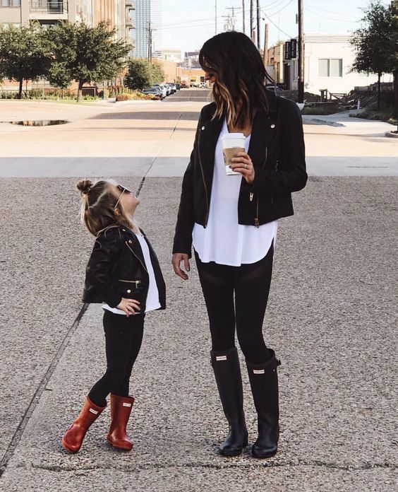 Madre e hija pequeña posando en la calle mientras visten pantalón de mezclilla negro, blusas blancas, chaquetas de cuero negras y la niña calza botas de lluvia rojas y tiene lentes de sol oscuros y la madre calza botas de lluvia negras mientras sostiene un café de starbucks