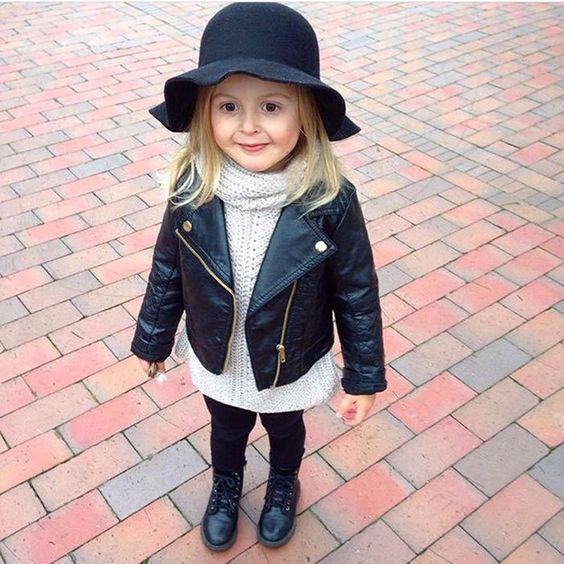 Niña rubia de cabello largo y suelto con sombrero negro, chaqueta de cuero negra, suéter blanco y botas negras altas