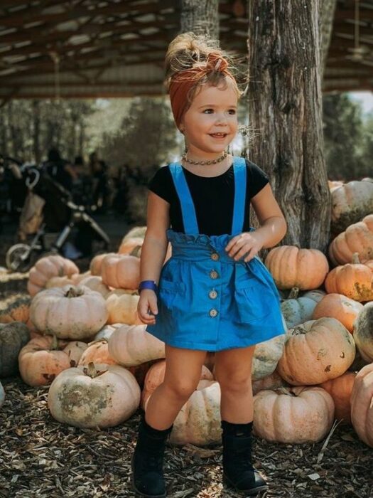 Niña rubia con banda naranja en la cabeza con blusa negra y falda de mezclilla y de fondo unas calabazas