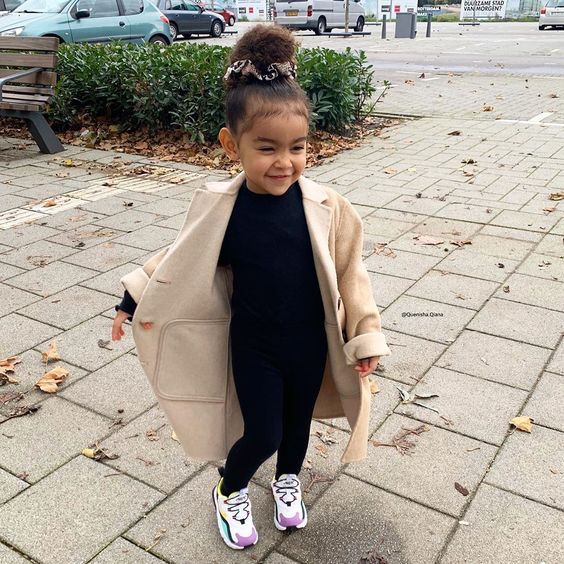 Niña pequeña de cabello rizado peinado en un chongo alto con blusa y pantalón negros, tenis blancos y abrigo beige