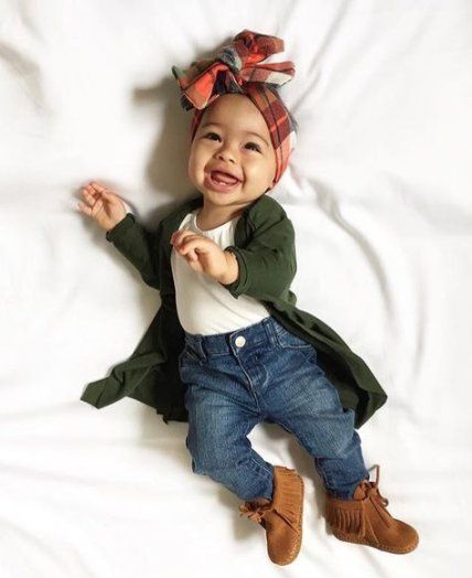 Bebé sonriente acostada en la cama con banda de colores en la cabeza, blusita blanca, cardigan verde esmeralda y pantalón de mezclilla