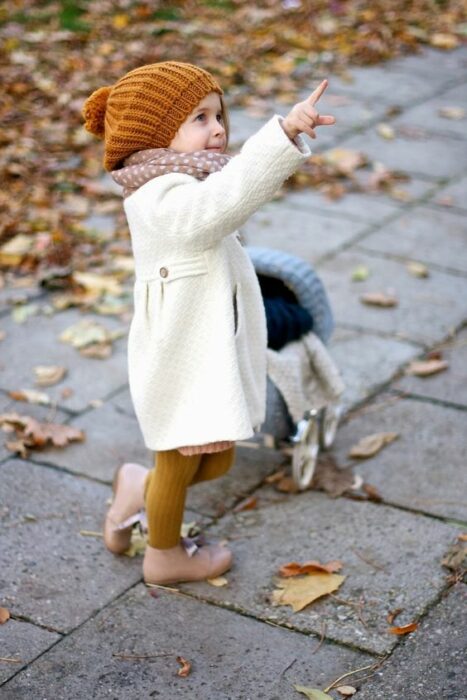Niña pequeña vistiendo abrigo blanco, gorrito naranja, mallas amarillo mostaza y botas beige