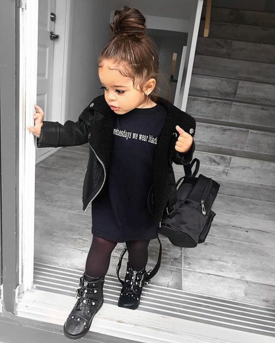 Niña pequeña con chonguito alto y blusa, chamarra, mallas y botas de color negro con mochilita negra