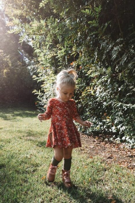 Niña rubia con coleta alta vistiendo vestido floreado color naranja con calcetas negras y botas cafés