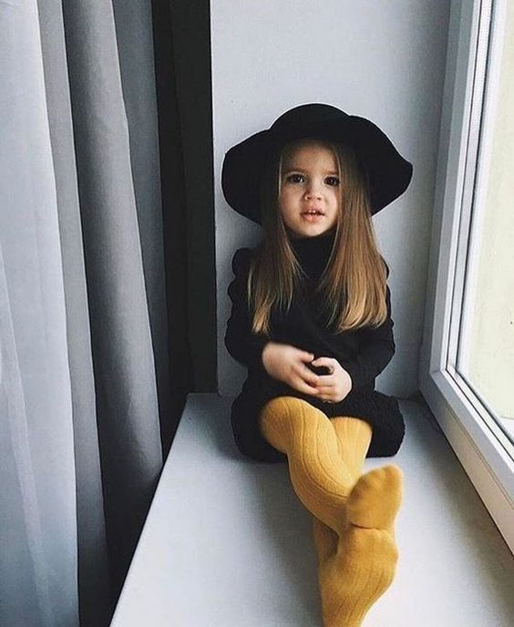 Niña de cabello castaño largo y liso sentada en una ventana con las piernas extendidas mientras viste un vestido negro, mallas amarillas y sombrero negro