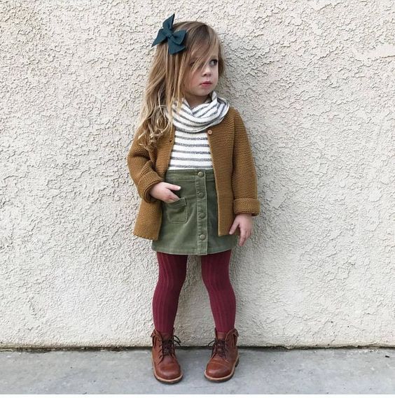 Niña rubia pequeña con cabello suelto sujetándose con un moño azul y vistiendo blusa blanca de rayas negras, suéter cagé, falda verde militar y medias guindas