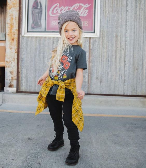 Niña rubia de cabello largo con gorrito gris vistiendo blusa gris, jeans y botas negras y una camisa amarilla amarrada a la cadera