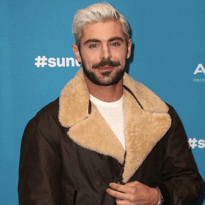 zac efron con el cabello teñido platinado, usando camiseta blanca y chamarra café oscuro de piel con cuello y solapas afelpadas