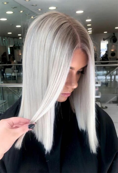 Chica con bata de estética negra mirando hacia abajo con melena lisa y raya en medio, con cabello blanco 'icy blonde'