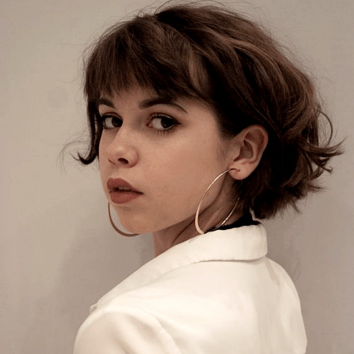 chica de cabello castaño con corte de cabello estilo bob francés, fleco corto, aretes dorados y blusa blanca de cuello formal