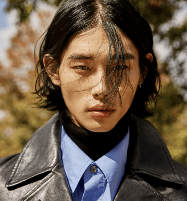 Tae Min Park; chico de cabello castaño usando un top negro de cuello alto, camisa azul de vestir y gabardina gruesa de cuero