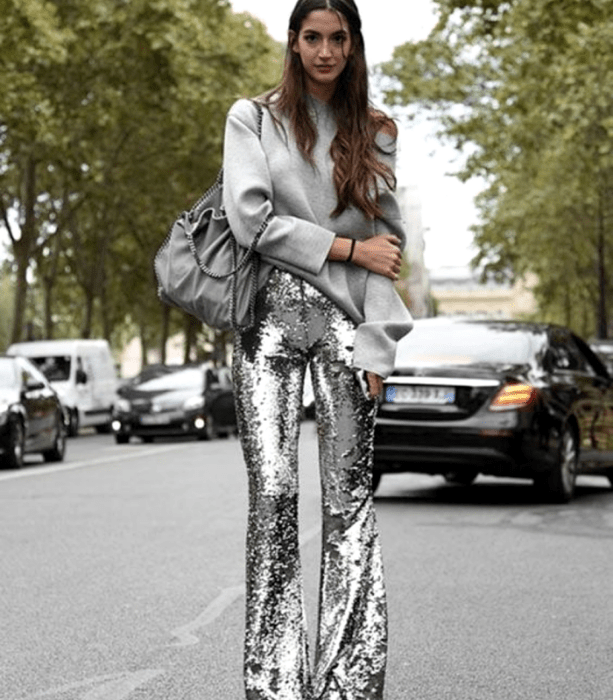 chica de cabello castaño con un suéter gris oversized, bolso gris, pantalones acampanados metálicos plateados