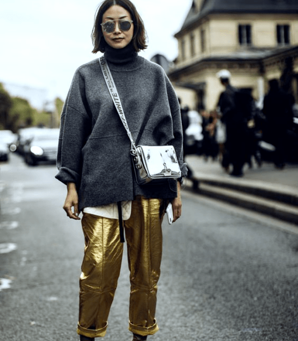 chica de cabello castaño con lentes de sol, sudadera gris de cuello alto, bolso metálico plateado, pantalones dorados metálicos