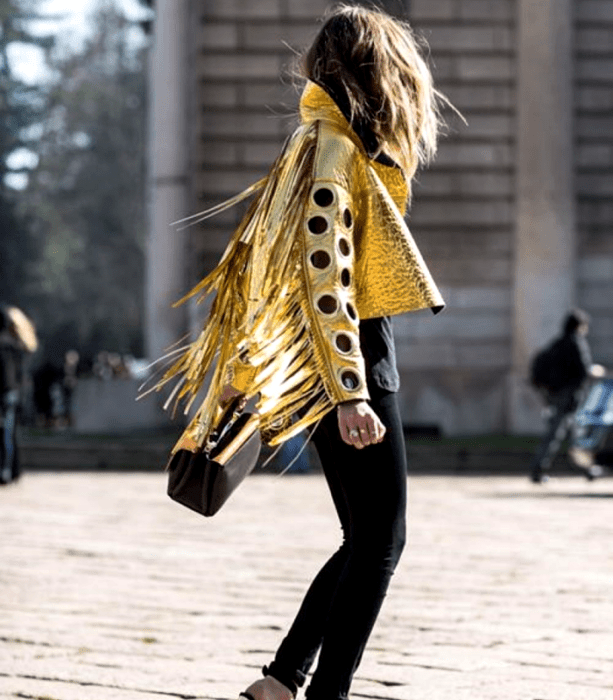 chica de cabello castaño usando una chaqueta dorada metálica de manga larga con perforaciones, skinny jeans negros, top negro y bolso de mano negro