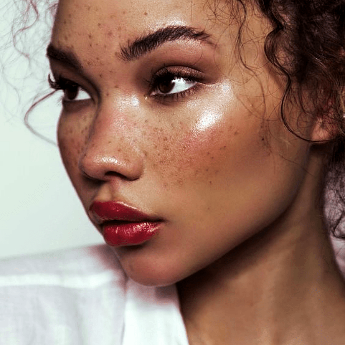 chica morena con cabello chino usando maquillaje sobre sus pecas naturales, lipstick rojo, blush rosa y sombras rosas