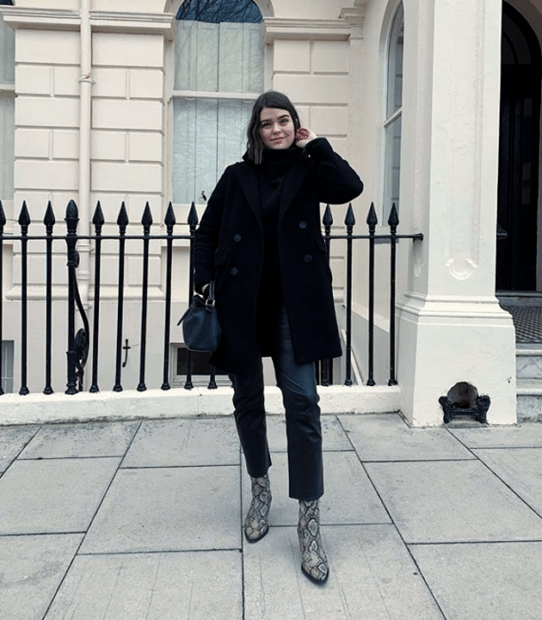 chica de cabello corto oscuro usando un abrigo negro, top negro de cuello alto y jeans con botines de piel y bolso negro
