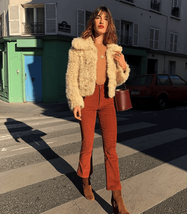 chica de cabello largo castaño usando un abrigo de peluche blanco con top naranja y pantalones rojos y botines