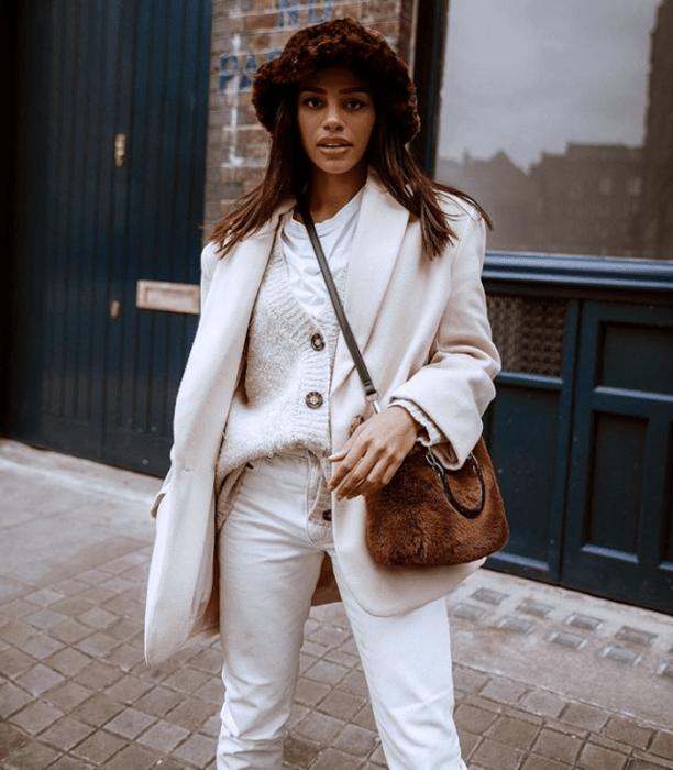 chica morena usando un bucket hat de piel con estampado animal print, abrigo beige, suéter blanco y pantalones blancos
