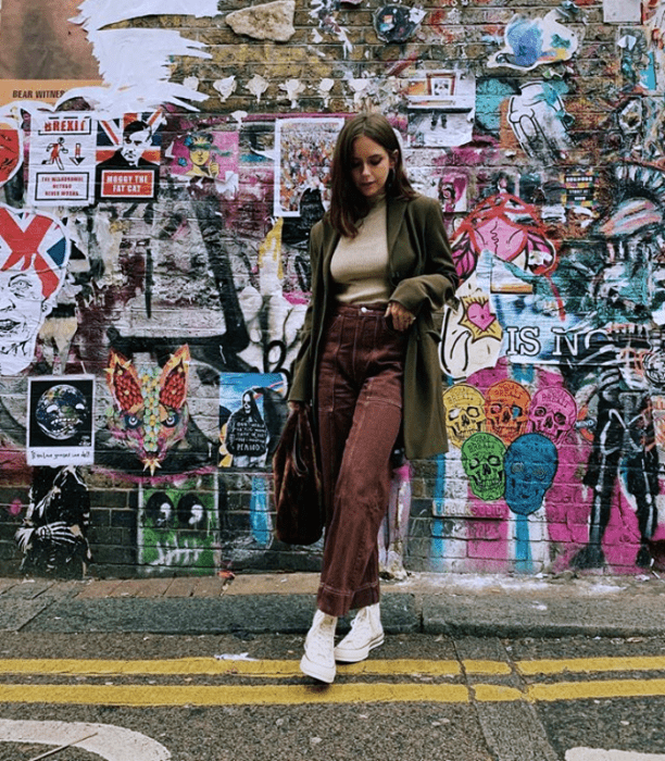 chica de cabello largo castaño usando un top beige, abrigo verde, pantalones marrones y tenis blancos