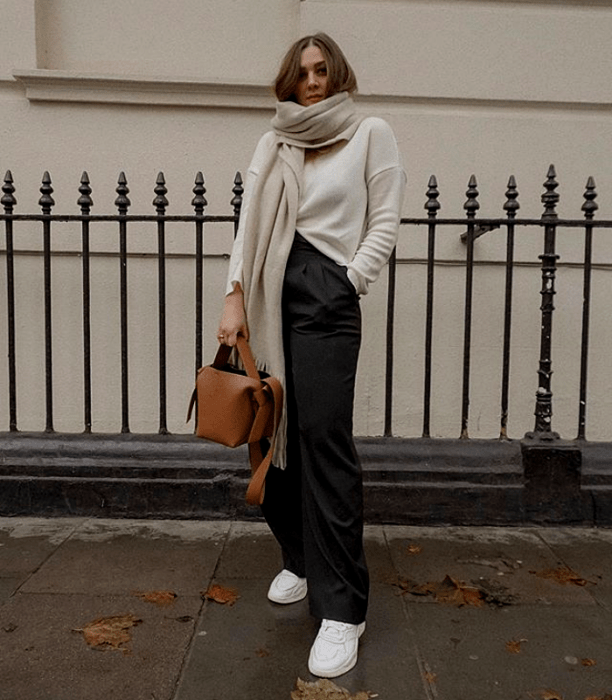 chica rubia usando una bufanda, suéter blanco, pantalones de vestir oscuros y tenis blancos con bolso café
