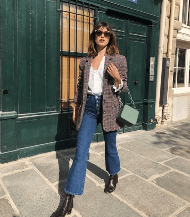 chica de cabello castaño largo usando lentes de sol, camisa blanca, chaqueta de cuadros, jeans acampanados y botines cafés