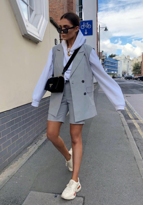Looks con sudadera; mujer caminando en la calle con lentes de sol, vestida con hoodie azul cielo, chaleco y short gris, tenis deportivos beige, bolsa negra