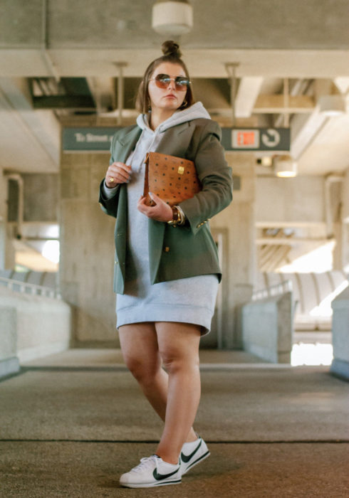 Looks con sudadera; chica plus size usando hoodie gris como vestido, con saco verde militar, tenis Nike blancos y bolsa de cartera café; peinado para cabello corto con medio chongo alto
